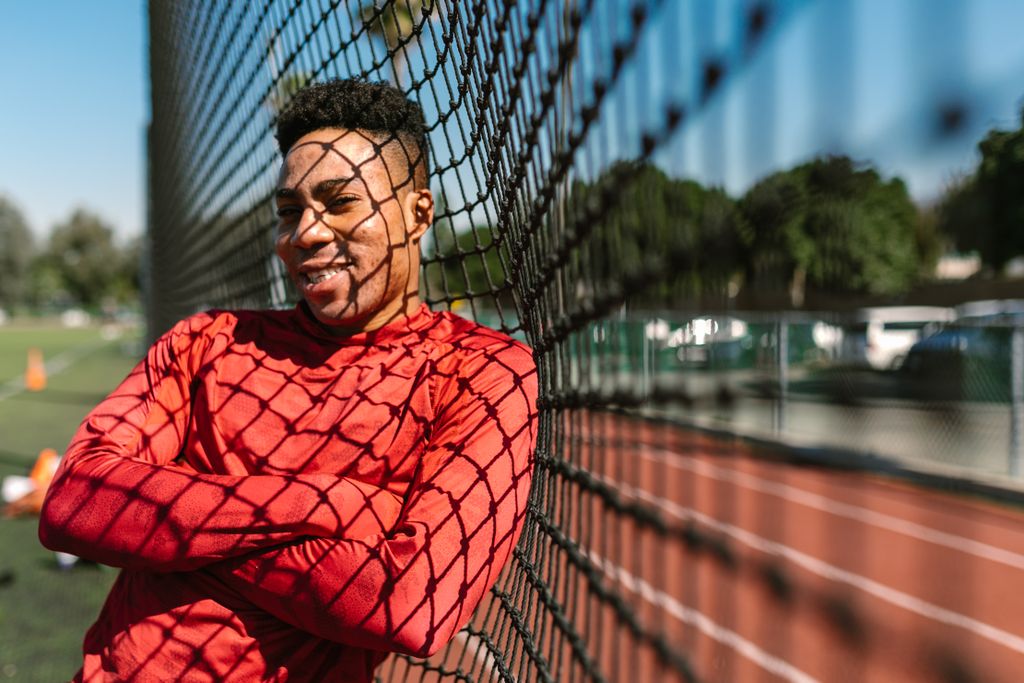 A cheerful athlete in a red shirt leaning on a sports field fence on a sunny day.